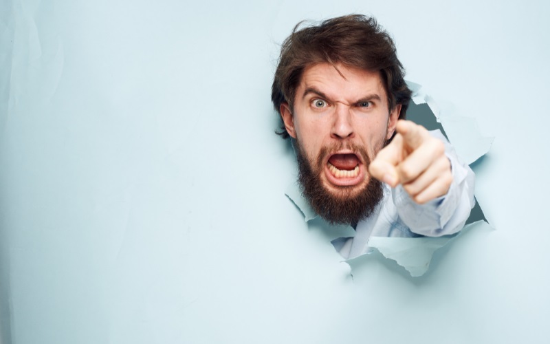 An angry man pointing his finger appearing to burst through a wall made of paper